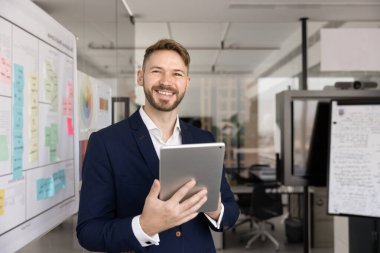 İş için internet teknolojisini tablette kullanan başarılı genç bir yönetici, ofis tahtasında dijital bir cihaz tutuyor, kameraya bakıyor, gülümsüyor, profesyonel iş portresi için.