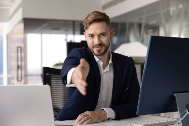 Kendine güveni tam genç bir iş adamı el sıkışırken kameraya, çalışma masasında bilgisayarla, laptopla, monitörle, işbirliği öneriyor. Erkek iş portresi