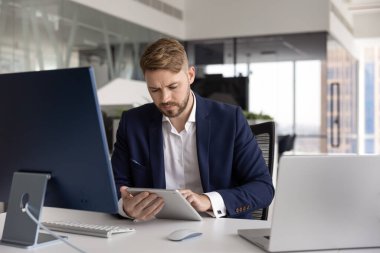 Ciddi genç bir finans tüccarı dijital tablet üzerinde ticaret uygulaması kullanıyor, bir çok cihazla ofis masasında oturuyor, laptop, bilgisayar monitörü, ped üzerinde daktilo, web servisi ile çalışıyor.