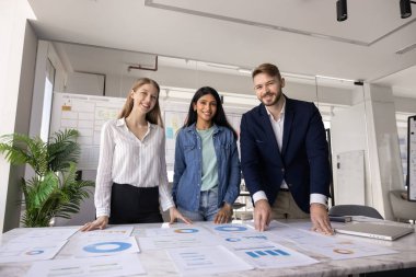 Pazarlama ajansı genç çalışanları büyük çalışma masasında kağıt raporları, şemalar, çizelgeler, kameraya bakıp gülümsüyorlar. Çeşitli şirket çalışanlarının takım portresi