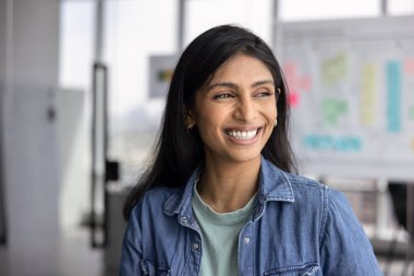 Neşeli genç Hintli genç iş kadını ofiste portre için poz veren, dişlek gülümsemeli, gülen, başarılı bir proje üzerinde düşünen, profesyonel başarı.