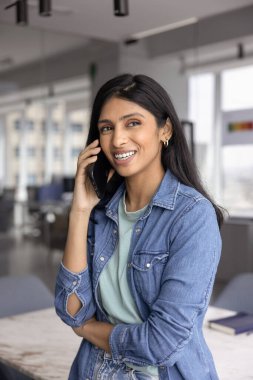 Mutlu genç Hintli şirket çalışanı cep telefonuyla konuşuyor, ofiste dikiliyor, başka tarafa bakıyor, gülümsüyor, telefon görüşmesinden zevk alıyor, iletişim kuruyor, gülümsüyor, dikey çekim yapıyor.