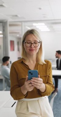 Ofiste elinde akıllı bir telefonla durup neşeli bir ifadeyle kameraya bakan orta yaşlı, gülümseyen profesyonel bir kadın. Yeni yapay zeka iş platformu, iş yerinde mobil uygulama kullanımı, dikey çekim