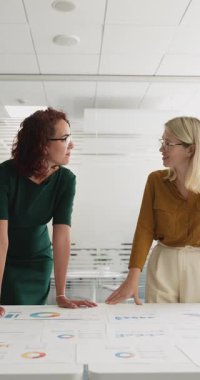 Modern ofiste proje üzerinde işbirliği yapan iki orta yaşlı profesyonel kadın, basılı belgeyi inceleyerek, spesifik ayrıntıları veya içgörüleri, analitik çizelgeleri ve grafikleri masaya yayarak,