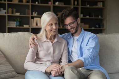 Anne ellerinin sıcaklığı. Sevecen gülümseyen yaşlı anne, kanepeye otur ve yetişkin oğluna sarıl. Sevgili yetişkin çocuğun kucaklamalarının tadını çıkar. Nazikçe elini tut.