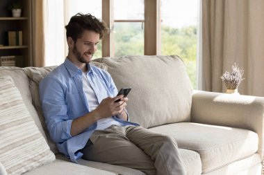 Bağlantıda kalmak. Genç hippi erkek girişimci modern konforlu apartman dairesindeki yumuşak kanepede dinleniyor. Sosyal medya sohbet uygulamasında cep telefonu mesajları tutuyor. Gülümse.