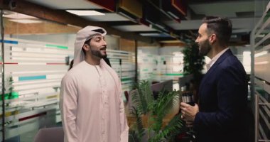 Geleneksel beyaz tobe giymiş genç bir Arap adam İspanyol iş arkadaşı ya da iş ortağıyla konuşuyor, el sıkışıyor, saygılarını sunuyor, iş yerinde toplantının sonucunu belirtiyor.