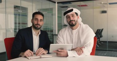 Çok uluslu iki adamın portresi modern ofis masasında elinde not defteri ve tablet cihazıyla kameraya poz vererek işbirliği içinde oturuyorlar. Kültürel çeşitlilik, takım çalışması ve stratejik planlama