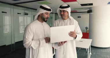 İki Arap iş adamı, geleneksel beyaz toblar giymiş, yeni iş platformlarını gözden geçirmek için modern yönetim kurulunda dizüstü bilgisayar kullanıyorlar, sunumları, borsa verilerini ve ticari detayları tartışıyorlar.