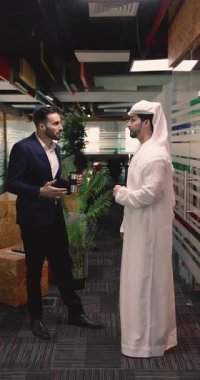 Modern bir iş yerinde, biri takım elbiseli, diğeri geleneksel Orta Doğulu iki iş adamı, etkileşim içinde, kültürler arası işbirliğini ve iş iletişimini sembolize ediyor.