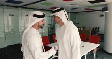 Geleneksel Orta Doğu tarzı giyinmiş iki Arap erkeği sıcak bir şekilde el sıkışarak birbirlerini selamlıyor ve ardından saygı dolu bir yanak öpücüğü, karşılama ve kültür geleneği olarak ofiste duruyorlar.