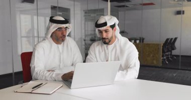 Geleneksel beyaz kanduras iki Arap işadamı modern ofiste konferans masasında oturmuş iş teklifini tartışıyor, dikkatle dizüstü ekrana bakıyor, verileri veya dijital sunumu inceliyor