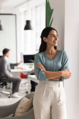 Güzel, tatmin olmuş Latin iş kadını, modern iş yerinde kendine güvenen bir çalışan mutlu bir gülümsemeyle mesafeye bakıyor, fikir üzerinde kafa yoruyor, iş yerinde yaratıcı çözümler arıyor.