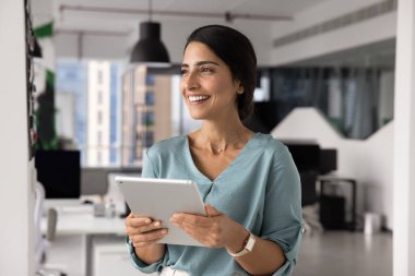 Modern ofiste dijital tablet cihazıyla duran mutlu düşünceli Latin iş kadını mutlu bir gülümsemeyle dikkati dağılmış, yapay zeka platformu ya da kurumsal uygulama kullanarak uzaktan iş akışının tadını çıkarıyor.