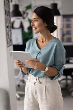 Latin iş kadını gündelik kıyafetleriyle modern ofiste duruyor. Tableti derin düşüncelere dalmış, tatmin olmuş bir ifadeyle uzaklığa bakıyor, dikkati dağılmış bir şekilde çevrimiçi görev tamamlıyor, fikirler ya da çözümler üzerinde kafa yoruyor.