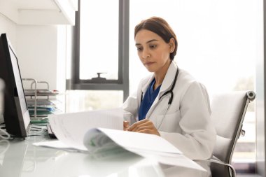 Laboratuvar önlüklü ve steteskoplu genç Ortadoğulu kadın hasta kayıtlarını okumakla meşgul. Klinikte tıbbî geçmişi olan, çalışma masasında çalışan bir doktor.