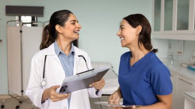 Klinik muayene odasında konuşan, sohbet eden, gülen, dosya tutan, alet tutan, takım çalışmasından, işbirliğinden, arkadaşlıktan keyif alan iki farklı doktor arkadaş.