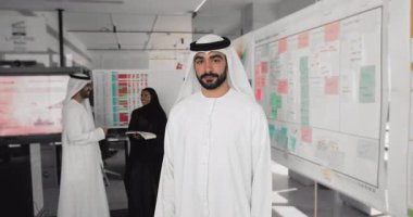 Beyaz Müslüman elbiseli, kendine güvenen yakışıklı genç Arap yönetici, çalışan iş adamlarıyla birlikte poz veriyor, güven jesti yapıyor, mutlu oluyor, kameraya bakıyor.