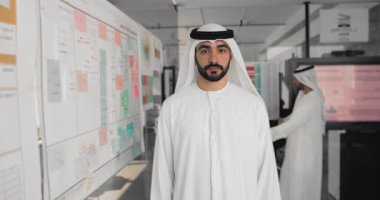 Arap bir adamın portresi, yönetici, beyaz kandora 'da proje yöneticisi modern işyerindeki planlama tahtalarının yanında duran, arka planda konuşan iki iş arkadaşı. Liderlik, kurumsal başarı, yönetim