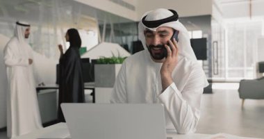 Geleneksel kandura ve ghutra 'da oturan Arap adam, dizüstü bilgisayarla uğraşırken telefonla ilgileniyor, iş yerinde olumlu iş iletişimini yönetiyor. Modern teknolojiler ve uzak işbirliği