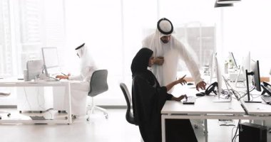 Modern çalışma ofisinde işbirliği içinde çalışan Arap profesyoneller, siyah tesettürlü kadın masasında otururken, beyaz kandura giymiş erkek meslektaşı görev ayrıntılarını, yeni proje ve yazılımı anlatıyor