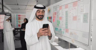 Genç Arap iş adamı, beyaz bulaşık makinesi giymiş ofis çalışanı iş yerinde akıllı telefon kullanıyor, mesajları paylaşıyor, e-postayla cevap veriyor, gülümsüyor, uzaktan dijital iş iletişiminden tatmin oluyor.