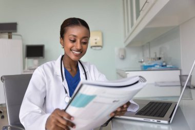 Beyaz üniformalı genç Afrikalı kadın doktor klinik muayenehanesinin modern muayene odasında masa başında oturuyor test sonuçlarını gözden geçiriyor, etkili bir tedavi ve hastanın iyileşme sürecinden tatmin oluyor.