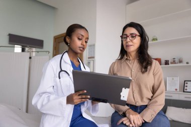 Palto giyen genç Afrikalı kadın pratisyen hekim, profesyonel klinik konsültasyon sırasında yaşlı kadın hastaya bilgi veriyor. Tıbbi açıklama, tanı incelemesi, tedavi planlaması