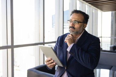 Dijital tablet taşıyan Hintli işadamı, düşünceli bir ifadeyle pencereden dışarı bakar, planlama stratejisi, karar verir, gelecek fırsatları araştırır, iş büyümesi için yeni olanaklar üzerine düşünür.