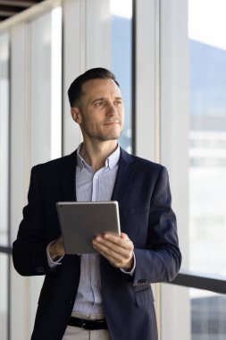 Düşünceli işadamı ofis penceresinin yanında duruyor, elinde dijital tabletle uzaklığa bakıyor, gelecekteki iş fırsatlarını değerlendiriyor, verileri analiz ediyor, yeni projeleri düşünüyor. Vizyon ve hırs