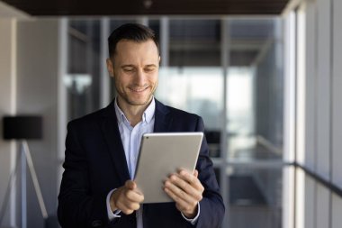 Resmi ceketli yakışıklı İspanyol işadamı ofis koridorunda modern dijital tablet cihazını kullanıyor, yazışmaları kontrol ediyor, sunum üzerinde çalışıyor, pazarlama stratejisi geliştiriyor. İş araçları kullanımı
