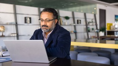 Ofis masasında oturan düşünceli işadamı, odaklanmış veya endişeli ifadeyle dizüstü bilgisayar ekranına bakıyor, rapor okuyor, proje detaylarını analiz ediyor, müşteri veya meslektaşlarına önemli e-postalar yazıyor.