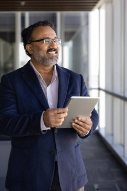 Gülümseyen yaşlı 45 'lik Hintli işadamı, şirket patronu veya CEO' su dijital tablet tutuyor, düşünceli bir ifadeyle pencereden dışarı bakıyor, liderliği ve profesyonelliği yayıyor, yenilikçi iş teknolojisi kullanımından keyif alıyor