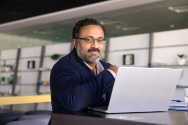 Masada oturan Hintli işadamının ofis portresi, dizüstü bilgisayarla çalışırken kameraya ciddi bir ifadeyle bak, görevleri yönet, e-postaları yönet, raporları analiz et, yaklaşan toplantıya hazırlanma