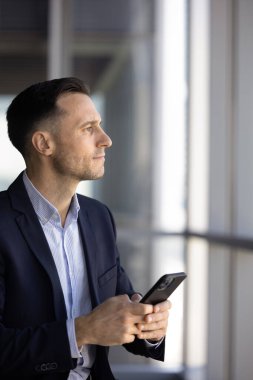 Orta yaşlı 40 'lı yılların iş adamı, modern akıllı telefonu ofisinde tutuyor, düşünceli bir ifadeyle, düşünceli bir şekilde uzaklardan bakıyor, önemli iş görüşmelerini bekliyor, yaklaşan fırsatları değerlendiriyor.
