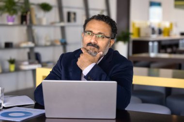 Düşünceli Hintli takım elbiseli işadamı dizüstü bilgisayarla masada oturur, derin stratejik düşünce, problem çözme, gelecekteki iş kararlarını planlarken, düşünceli bir ifadeyle mesafeye bakar.