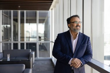 Orta yaşlı Hintli işadamı büyük ofis penceresinin önünde mutlu bir gülümsemeyle duruyor, yeni başlangıç fikirleri, gelecek fırsatları ve perspektifleri düşünüyor. İş görüşü, strateji oluşturma
