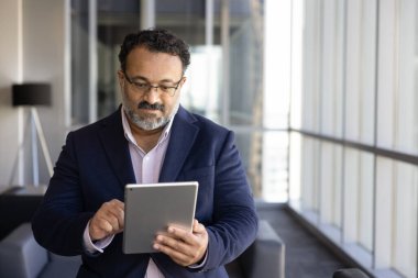 Takım elbiseli yetişkin Hintli işadamı ofis koridorunda duruyor, tablete odaklanıyor, kablosuz cihaz üzerinde çalışıyor, müşteriye e-posta ile cevap veriyor, pazarlama stratejisi hazırlıyor. Yenilik, dijital iletişim