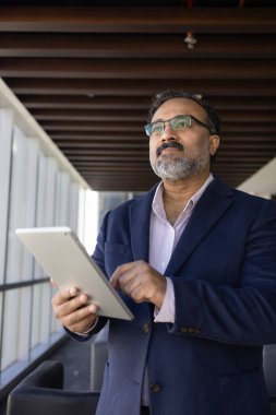 Olgun 45 'lik Hintli işadamı modern ofis koridorunda elinde dijital tabletle dikiliyor, odaklanmış, düşünceli bir ifadeyle, düşünceli fikirlerle, verileri gözden geçiriyor. Stratejik planlama, liderlik
