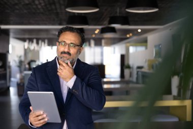 Takım elbiseli yaşlı, düşünceli Hintli bir adam iş yerinde çalışan tablet hakkında bilgi analiz ediyor, iş kararı üzerinde kafa yoruyor, ortakların önerilerini gözden geçiriyor ya da yenilikçi bir strateji düşünüyor.