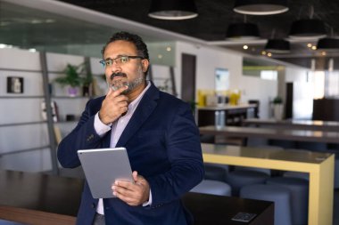 Resmi giyinmiş olgun iş adamı modern açık alan ofisinde düşünceli bir şekilde duruyor, tablet tutuyor, önemli proje güncellemesi üzerinde kafa yoruyor, yeni pazar eğilimlerini değerlendiriyor, yenilikçi fikirleri değerlendiriyor.
