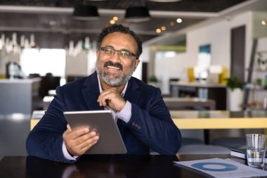 Mutlu Hintli işadamı tabletle aynı masada oturuyor, neşeli bir ifadeyle başka tarafa bakıyor, üretken bir iş akışı içinde, iyi iş haberleri alıyor, yenilikçi yapay zeka teknolojisinin tadını çıkarıyor.