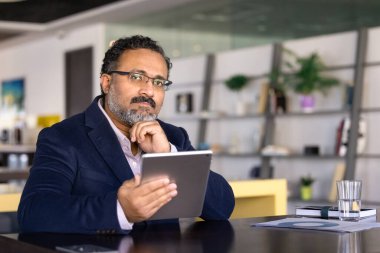 Ciddi yaşlı Hintli işadamı elinde tabletle modern ofis masasında oturmuş, cihazla ilgili mali raporları ya da stratejik verileri analiz ediyor, gelecekteki iş stratejilerini ya da kararlarını düşünüyor,