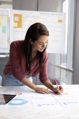 Odaklanmış Latin iş kadını notlar alıyor, yapışkan kağıda önemli noktalar yazıyor, analiz ediyor, diyagramlar, pasta ve bar grafiklerinde gösterilen verileri düzenliyor. Araştırma, analitik iş akışı