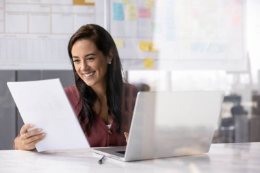 Gazetelerde verileri inceleyen gülümseyen kadın dizüstü bilgisayarla masada oturuyor. Ofis çalışanı mali raporları analiz eder, sözleşmeleri gözden geçirir, piyasa araştırması yapar, yatırım fırsatlarını değerlendirir.