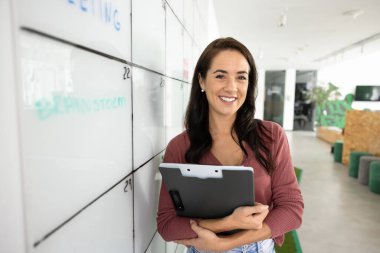 Profesyonel kadın özgüvenle gülümsüyor, el yazısıyla yazılmış notlarla büyük bir tahtanın yanında duruyor, kameraya bakıyor, modern ofisteki görevleri planlamak, düzenlemek veya yönetmekle meşgul bir pano tutuyor.