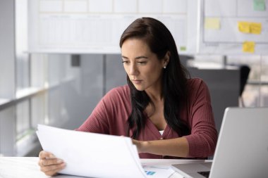 İşyerinde belgelerle çalışan, elinde kağıtlar tutan, sözleşmeleri ya da teklifleri inceleyen, mali raporları analiz eden, iş verilerini çizelge ve diyagramlarda inceleyen, stratejik kararlar alan odaklı bir kadın.