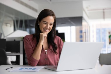 İş yerinde dizüstü bilgisayarda çalışan gülümseyen bir kadın, analitik verileri olumlu sonuçlarla gözden geçirir, satış artışından tatmin olur, iyi sonuç verir, ara sırasında bilgisayardan eğlenceli çevrimiçi içeriği izler.
