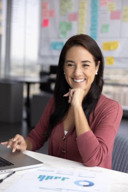 Ofiste dizüstü bilgisayarla masada oturan gülümseyen profesyonel kadın işten memnun, renkli grafik ve çizelgeleri olan belgeler yakınlarda. Veri analizi, başarılı sonuç, analiz