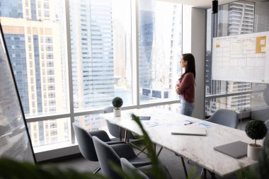 Kurumsal vizyon ve stratejik yansıma. Düşünceli profesyonel kadın büyük panoramik pencerenin yanında duruyor, büyük şehir siluetine bakıyor, derin düşünce anı, gelecek planlama, iş perspektifleri